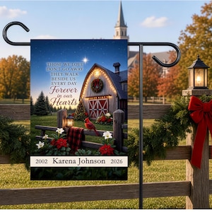May include: A rectangular garden flag featuring a barn scene with a red cardinal, poinsettias, and a plaid blanket on a fence. The text reads: Those we love don't go away they walk beside us every day forever in our hearts, 2002 Karena Johnson 2026.