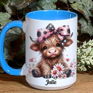 A beautifully designed personalized Highland Cow mug featuring an adorable brown cow with a pink and black polka dot bow, surrounded by soft pink flowers. This 15-ounce ceramic mug is available with a black, white, or blue handle.