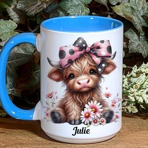 A beautifully designed personalized Highland Cow mug featuring an adorable brown cow with a pink and black polka dot bow, surrounded by soft pink flowers. This 15-ounce ceramic mug is available with a black, white, or blue handle.