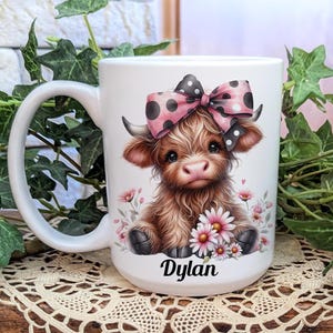 A beautifully designed personalized Highland Cow mug featuring an adorable brown cow with a pink and black polka dot bow, surrounded by soft pink flowers. This 15-ounce ceramic mug is available with a black, white, or blue handle.