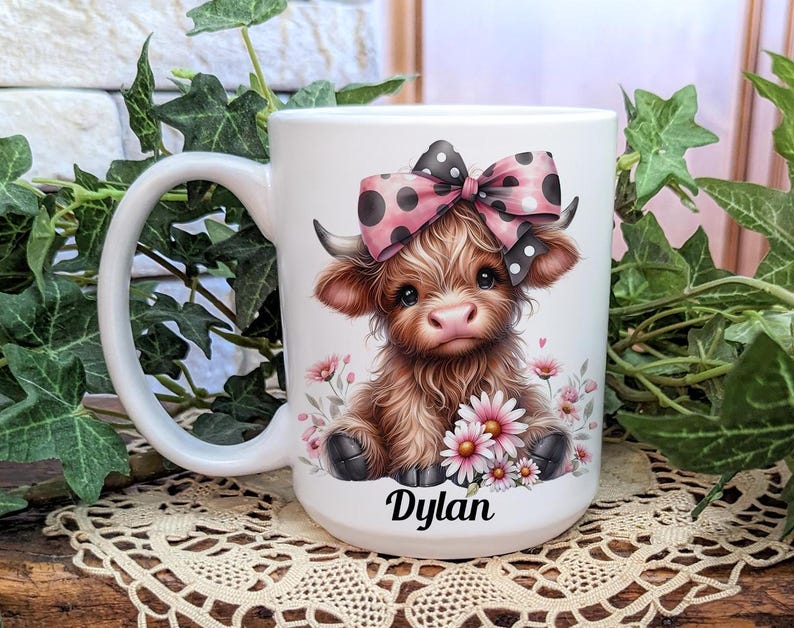 A beautifully designed personalized Highland Cow mug featuring an adorable brown cow with a pink and black polka dot bow, surrounded by soft pink flowers. This 15-ounce ceramic mug is available with a black, white, or blue handle.