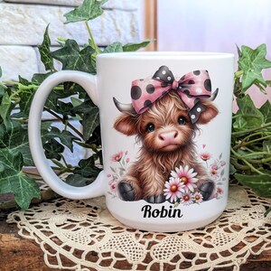 A beautifully designed personalized Highland Cow mug featuring an adorable brown cow with a pink and black polka dot bow, surrounded by soft pink flowers. This 15-ounce ceramic mug is available with a black, white, or blue handle.