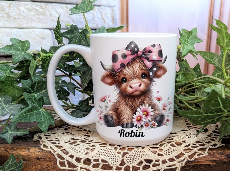 A beautifully designed personalized Highland Cow mug featuring an adorable brown cow with a pink and black polka dot bow, surrounded by soft pink flowers. This 15-ounce ceramic mug is available with a black, white, or blue handle.