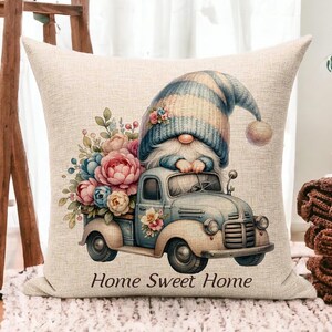 May include: A decorative pillow with a whimsical design. It features a gnome driving a vintage blue truck filled with flowers. The pillow has the text "Home Sweet Home" at the bottom. The gnome wears a blue and beige striped hat.