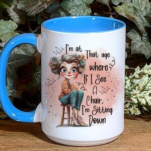 May include: White ceramic coffee mug with a blue rim and handle. The mug features a cartoon illustration of a woman with a whimsical expression and the text "I'm at that age where if I see a chair, I'm sitting down."