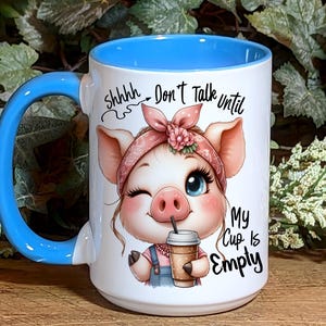 May include: White ceramic mug with a blue rim and handle. The mug features a cartoon pig wearing a pink bandana with flowers and holding a cup of coffee. The text on the mug reads "Shhhh... Don't Talk Until My Cup Is Empty."