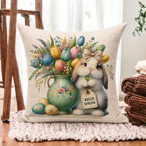 May include: A decorative square pillow with a spring theme. The pillow features a watercolor illustration of a bunny holding a tag that says "Hello Spring". A vase of colorful tulips and painted Easter eggs complete the design.