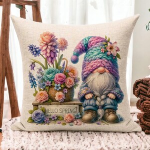 May include: A decorative pillow featuring a whimsical gnome with a pink and blue knitted hat, holding a basket of colorful flowers. The pillow has a white background with the text "Hello Spring!" printed on it.