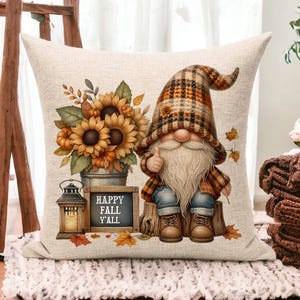 May include: A beige square throw pillow with a fall theme. It features a gnome with a long white beard, a plaid hat and shirt, and a thumbs-up gesture. A bouquet of sunflowers and the text "HAPPY FALL Y'ALL" are also on the pillow.