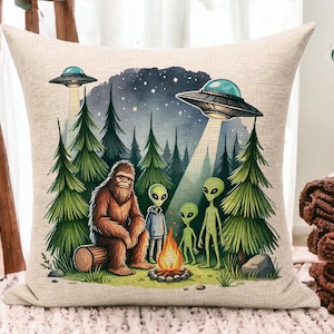 May include: A throw pillow with a whimsical illustration of a Bigfoot and three aliens in a forest setting. The aliens are wearing spacesuits and holding hands around a campfire. Two flying saucers are visible in the sky above the trees.