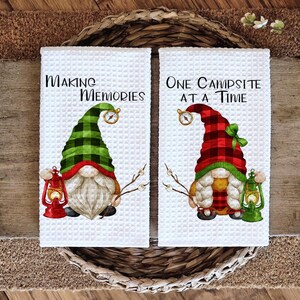 May include: Two white waffle-weave kitchen towels with gnome designs. One towel reads "Making Memories" and the other reads "One Campsite at a Time." Each gnome has a plaid hat, a lantern, and a compass.