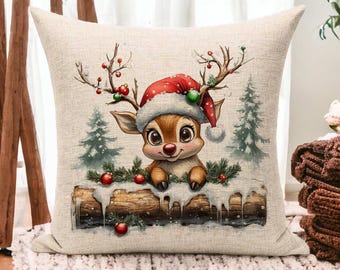 May include: A beige throw pillow with a Christmas-themed design featuring a cartoon reindeer wearing a red Santa hat and holding onto a log covered in snow. The reindeer is surrounded by green pine branches and red berries.