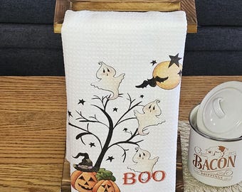 May include: A white kitchen towel with a Halloween theme featuring ghosts, a moon, stars, and a tree. The towel is hanging on a wooden towel rack with the word "BOO" printed on the towel.
