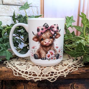 A beautifully designed personalized Highland Cow mug featuring an adorable brown cow with a pink and black polka dot bow, surrounded by soft pink flowers. This 15-ounce ceramic mug is available with a black, white, or blue handle.
