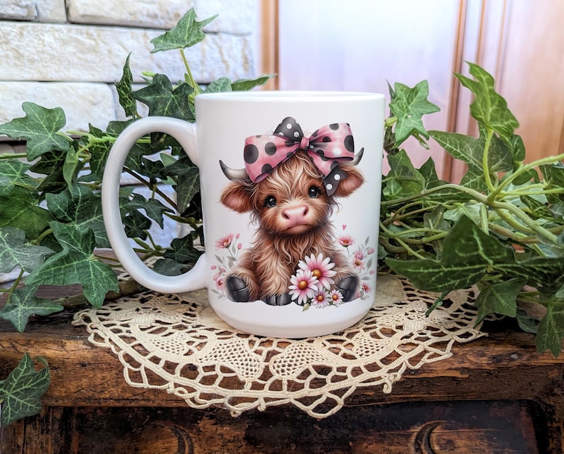 A beautifully designed personalized Highland Cow mug featuring an adorable brown cow with a pink and black polka dot bow, surrounded by soft pink flowers. This 15-ounce ceramic mug is available with a black, white, or blue handle.
