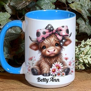 A beautifully designed personalized Highland Cow mug featuring an adorable brown cow with a pink and black polka dot bow, surrounded by soft pink flowers. This 15-ounce ceramic mug is available with a black, white, or blue handle.