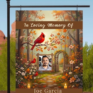 May include: A rectangular garden flag featuring the text In Loving Memory Of and Joe Garcia. The design displays a stone path leading to a wooden gate, surrounded by autumn trees, red cardinal birds, white flowers, and two lit candles.