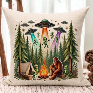 May include: A throw pillow with a whimsical illustration of a Bigfoot sitting by a campfire with aliens in the forest. The aliens are flying in UFOs and there are trees and a tent in the background.