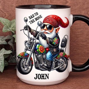 May include: White ceramic coffee mug with a black rim and handle. The mug features a cartoon illustration of a gnome riding a motorcycle with the text "Bad to the Bone" and the name "JOHN" printed on the mug.