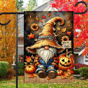 May include: A rectangular garden flag featuring a gnome wearing a large tan hat, a plaid shirt, and blue overalls. The gnome is surrounded by orange pumpkins, sunflowers, and autumn leaves against a dark background. A sign reads Welcome Fall.