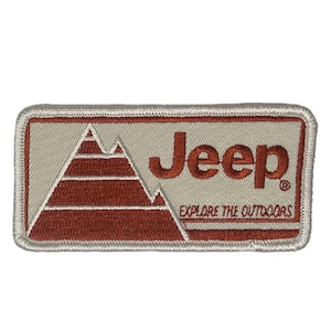 May include: Embroidered patch featuring the word "Jeep" in a rust-colored font, with a mountain graphic. The patch also includes the phrase "EXPLORE THE OUTDOORS" in a smaller font. The patch has a rectangular shape with a light gray background and a rust-colored border.