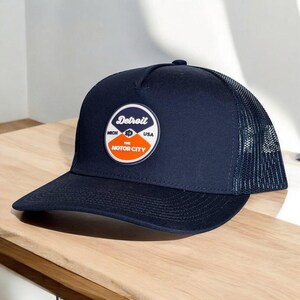 May include: Navy blue trucker hat with a mesh back and a curved brim. The hat features a circular patch with the word "Detroit" and the text "The Motor City" in white, blue, and orange.