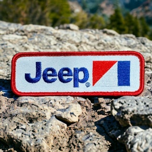 Puede incluir: Un parche rectangular blanco con costuras rojas y azules alrededor de los bordes. El parche presenta la palabra "Jeep" en bordado azul y un logotipo rojo, blanco y azul.