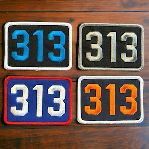 Puede incluir: Cuatro parches bordados con el número 313 en diferentes colores. Los parches son negros con costuras blancas, negros con costuras plateadas, azules con costuras blancas y negros con costuras naranjas.