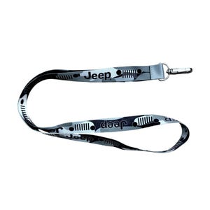 Könnte beinhalten: Ein grau-weißes Tarnmuster-Schlüsselband mit dem Wort "Jeep" in wiederholter Schrift. Das Schlüsselband hat einen silberfarbenen Metallclip zum Befestigen von Schlüsseln oder einem Ausweis. Das Design beinhaltet das Jeep-Logo.