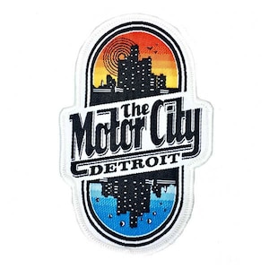 Op de afbeelding: Geborduurde patch met de tekst "The Motor City DETROIT". Het ontwerp toont een silhouet van een stadsgezicht tegen een zonsondergang, met een blauw-witte rand. De patch heeft een ovale vorm.