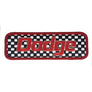 Op de afbeelding: Een rechthoekige patch met het woord "Dodge" in het rood, geborduurd over een zwart-wit geblokte achtergrond. De patch heeft een rode rand. Deze patch is waarschijnlijk voor autoliefhebbers.