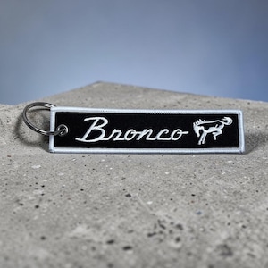 May include: Black rectangular keychain with white embroidered text "Bronco" and a horse silhouette. The keychain has a silver metal ring for attaching to keys. The edges are trimmed with white stitching.