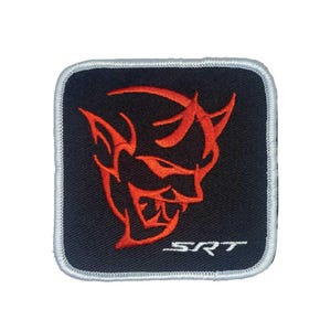 May include: Square black patch with a red devil face design and the letters "SRT" in white. The patch has a white border. The devil face has horns and a menacing expression. The patch is likely for a car or automotive enthusiast.
