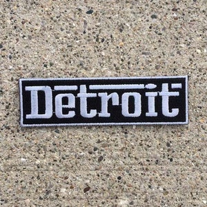 Könnte beinhalten: Ein rechteckiger Aufnäher mit dem Wort "Detroit" in weißen Blockbuchstaben auf schwarzem Grund. Der Aufnäher hat einen schwarzen Rand und liegt auf einer strukturierten, hellfarbigen Oberfläche.