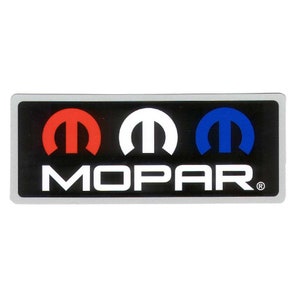 Puede incluir: Pegatina rectangular con el logotipo de Mopar. El logotipo presenta tres formas estilizadas de 'M' en rojo, blanco y azul, sobre la palabra 'MOPAR' en blanco, sobre un fondo negro con un borde plateado.