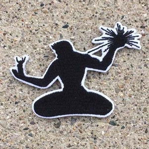 Könnte beinhalten: Schwarz-weißer, gestickter Aufnäher mit einer stilisierten Silhouette einer Person, die einen Lichtblitz hält. Der Aufnäher hat einen weißen Rand und befindet sich auf einer strukturierten, grauen Oberfläche. Das Design ist einfach und grafisch.