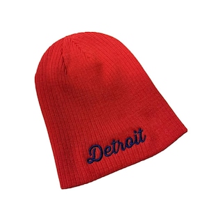 May include: A red ribbed knit beanie with the word "Detroit" embroidered in navy blue script. The hat is a classic style, suitable for cold weather and casual wear.