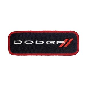 Puede incluir: Un parche rectangular con la palabra "DODGE" bordada en blanco sobre un fondo negro. El parche tiene un borde rojo y el logotipo de Dodge en rojo.