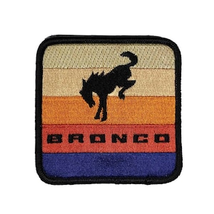 May include: An embroidered patch featuring a stylised horse head in black and white, with the word "BRONCO" in black text. The patch has a striped background with beige, orange, red and blue stripes.