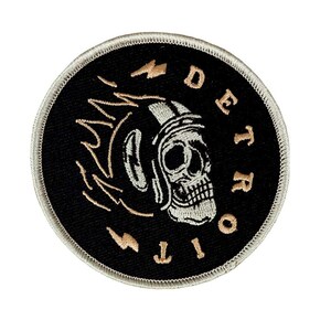 May include: Round black embroidered patch featuring a skull wearing a motorcycle helmet. The patch has the words "DETROIT CITY" in a circular pattern around the skull. The design includes tan lightning bolts and a silver border.