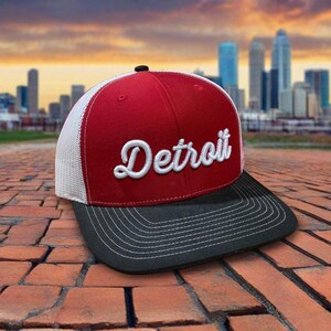 May include: A red and white Detroit baseball cap with a black brim. The word "Detroit" is embroidered in white script across the front. The cap has a mesh back and a black button on top.