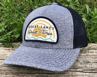 May include: A gray and black trucker hat with a patch that says "Great Lakes Great Times" in a retro font. The patch features a yellow sun with a blue wave underneath.