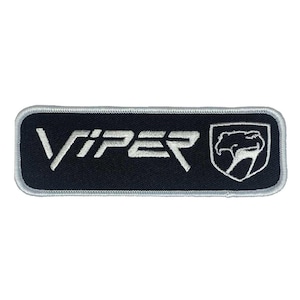 May include: A rectangular black patch featuring the word "VIPER" in white embroidery, accompanied by a white emblem of a snake's head. The patch is edged with silver.
