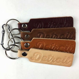 May include: Four leather keychains with a silver key ring. Each keychain has the word "Detroit" embossed in a different color: burgundy, brown, tan, and natural.