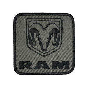 May include: A square, olive green patch with a black border. The patch features the RAM logo, a stylized ram's head, above the word "RAM" in bold, black capital letters. The patch has a textured appearance.