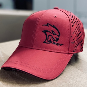 Può includere: Un cappello da baseball rosso con un logo ricamato nero di una bestia ringhiante e il testo "SRT". La parte posteriore del cappello ha un design a motivi bianco e nero.