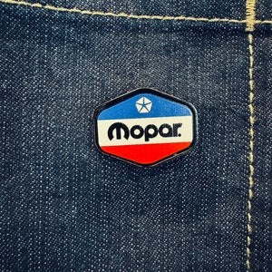 Puede incluir: Un parche hexagonal con el logotipo de Mopar en colores azul, blanco y rojo. El logotipo presenta una estrella de cinco puntas y la palabra "Mopar" en letras negras.