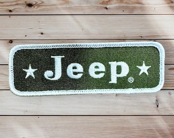 Op de afbeelding: Een groene stoffen patch met witte stiksels en het woord "Jeep" in witte letters met een witte ster aan elke kant.