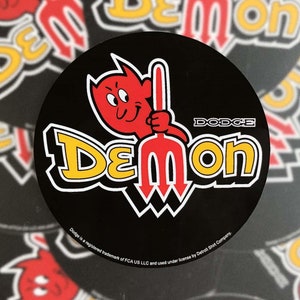 Puede incluir: Una pegatina redonda negra con un diablo rojo de dibujos animados sosteniendo un tridente. La palabra "Demon" está en amarillo y blanco, con "Dodge" encima. La pegatina tiene un borde blanco y texto en la parte inferior.