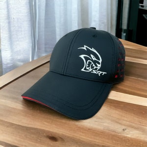 May include: Black baseball cap with a curved peak and a white embroidered logo featuring a stylized animal head and the letters "SRT." The cap has red accents on the peak and side panels. The cap is sitting on a wooden surface.